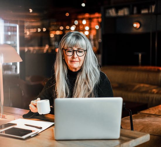 Kvinna med långt grått hår och glasögon arbetar på en laptop vid ett cafébord, medan hon håller en kaffekopp