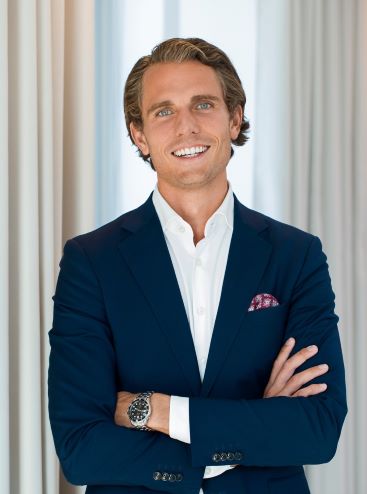 Man i mörkblå kostym med korslagda armar, står framför ljusa gardiner och ler mot kameran