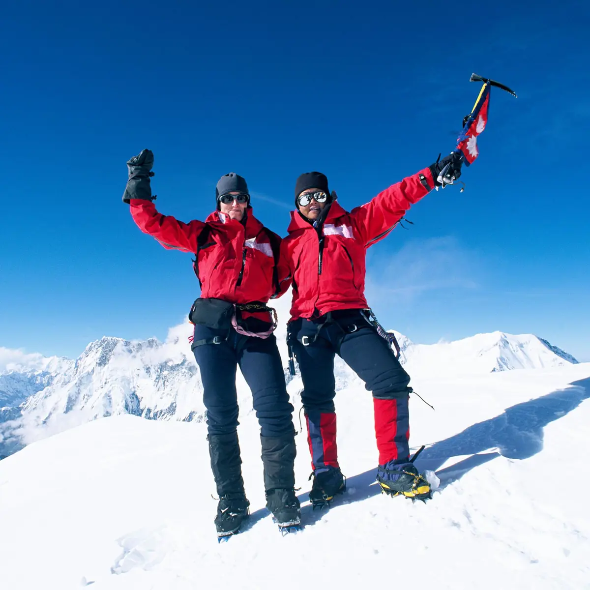 Två bergsbestigare i röda jackor firar på snötäckt bergstopp under klarblå himmel