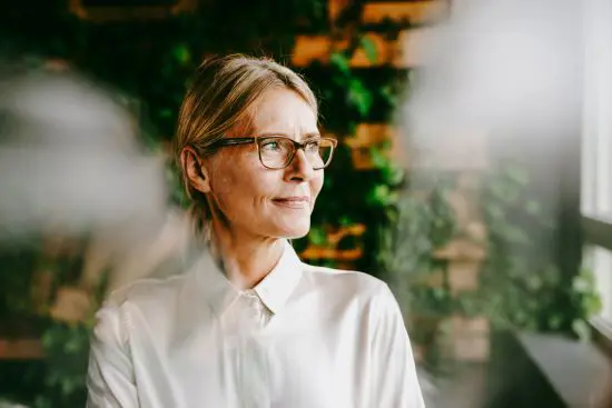 Kvinna med glasögon och vit skjorta tittar fundersamt ut genom fönstret, omgiven av gröna växter