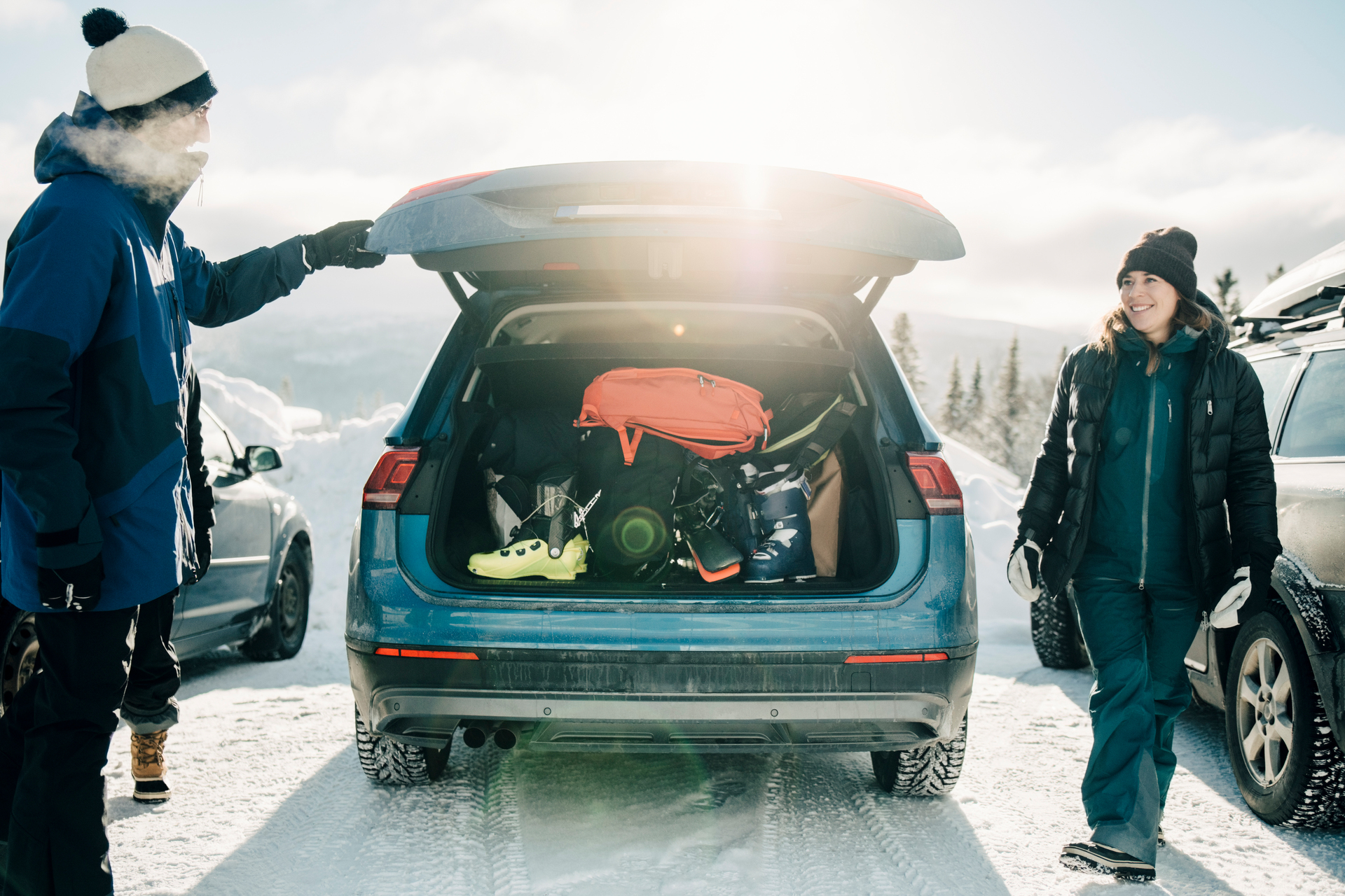 Två personer vid en bil parkerad i snön, med bagageutrymmet öppet och fyllt med skidutrustning, inklusive pjäxor och en ryggsäck, under en solig vinterdag