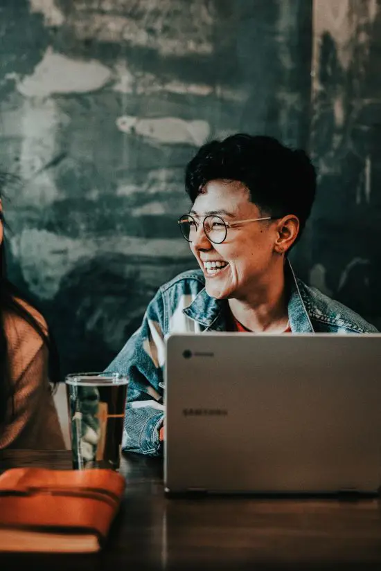 Person som ler och arbetar vid en bärbar dator i ett café, med ett glas dryck och en anteckningsbok på bordet