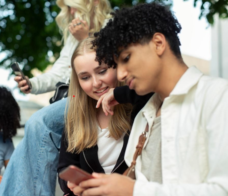 Ungdomar som använder smartphones utomhus, fokus på två personer som delar skärm och skrattar. Socialt umgänge och teknik