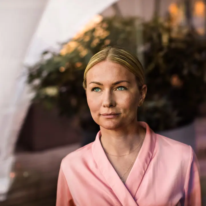 Kvinna i rosa blus framför suddig bakgrund, ser tankfull ut