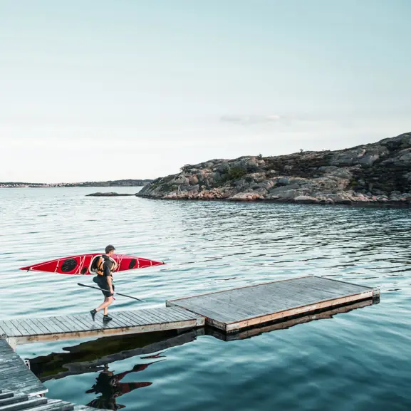 Person bär en röd kajak på en brygga vid en klippig kust, spegelblankt vatten och molnfri himmel i bakgrunden
