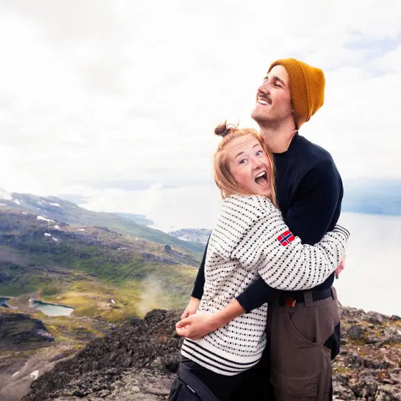 Par på en bergstopp omfamnar varandra glatt, med utsikt över en grönskande dal och sjöar i bakgrunden