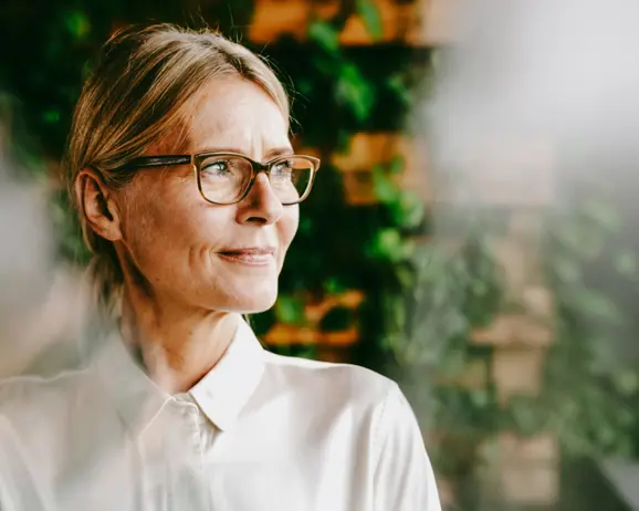 Äldre kvinna med glasögon och vit skjorta ler med blicken åt sidan, omgiven av grönska i bakgrunden