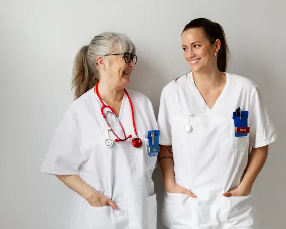 Två kvinnliga vårdpersonal i vita uniformer, en äldre och en yngre, står tillsammans och ler mot varandra med stetoskop och namnskyltar