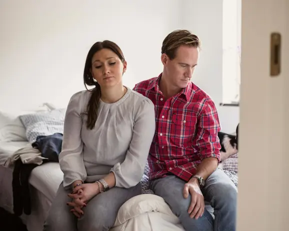 Par i avslappnade kläder sitter på sängkanten i ett sovrum, ser fundersamma ut