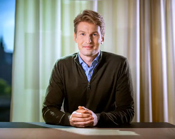 Man klädd i en mörk tröja sitter vid ett bord med händerna ihop, mot en bakgrund av gardiner och mjukt ljus