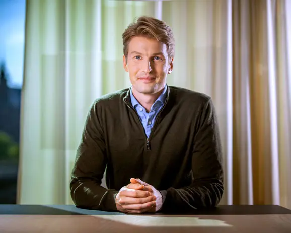 Man klädd i en mörk tröja sitter vid ett bord med händerna ihop, mot en bakgrund av gardiner och mjukt ljus