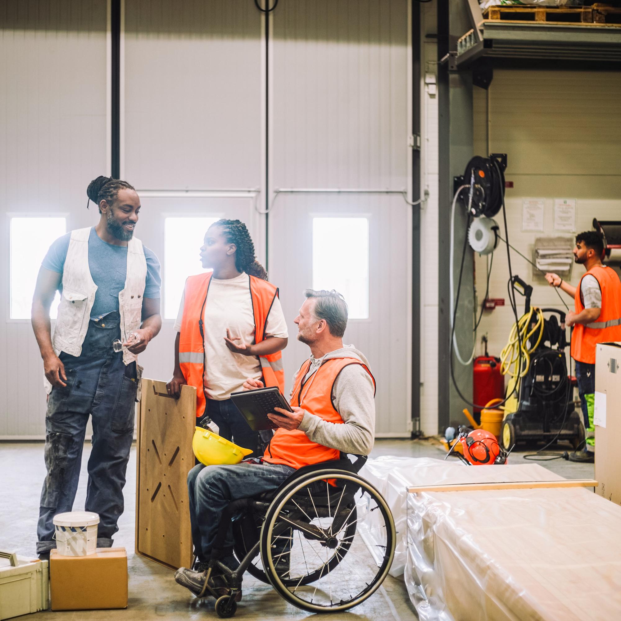 Arbetare i reflexvästar i en lagerlokal, samtalar och samarbetar. En person i rullstol diskuterar med två kollegor