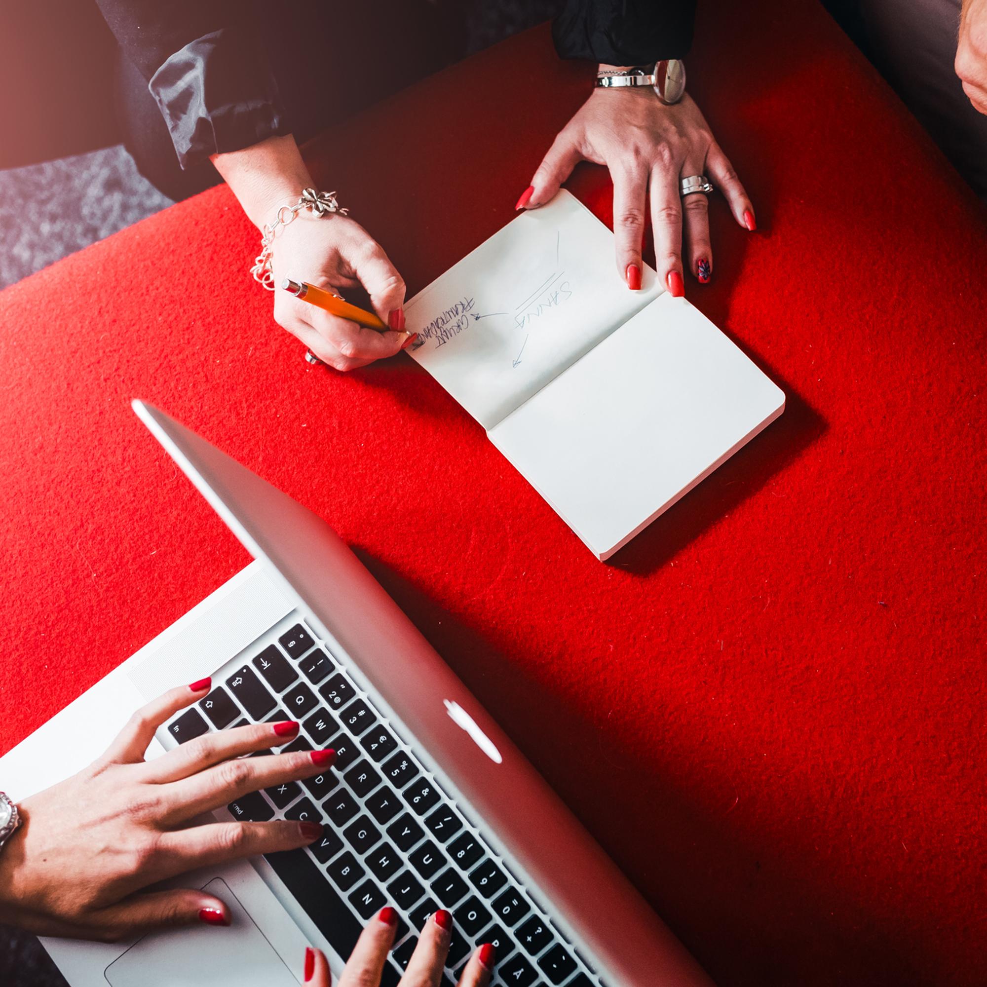 Person som skriver i anteckningsbok bredvid en bärbar dator på ett rött bord