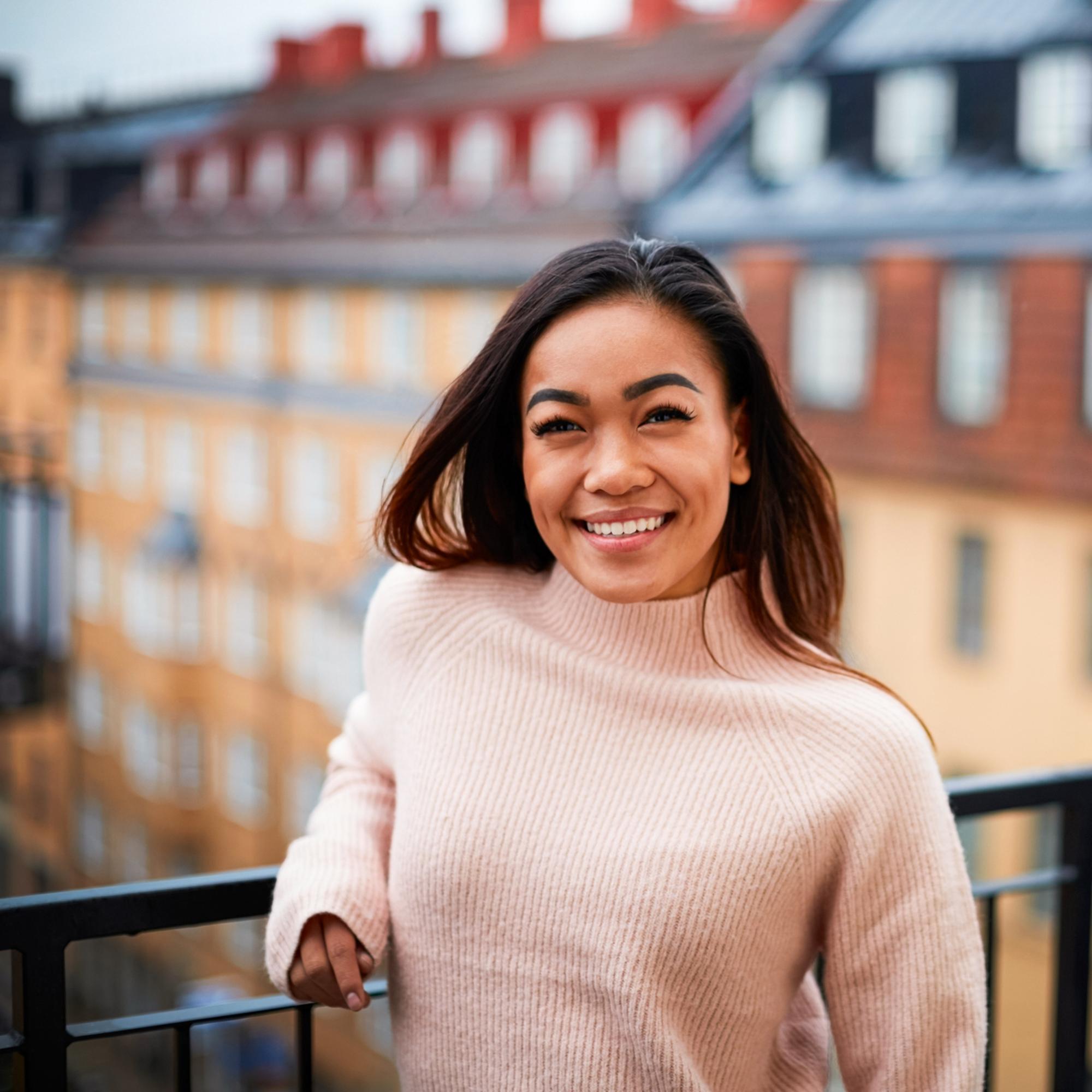 Leende kvinna i beige tröja på balkong med Stockholms stadsbild i bakgrunden