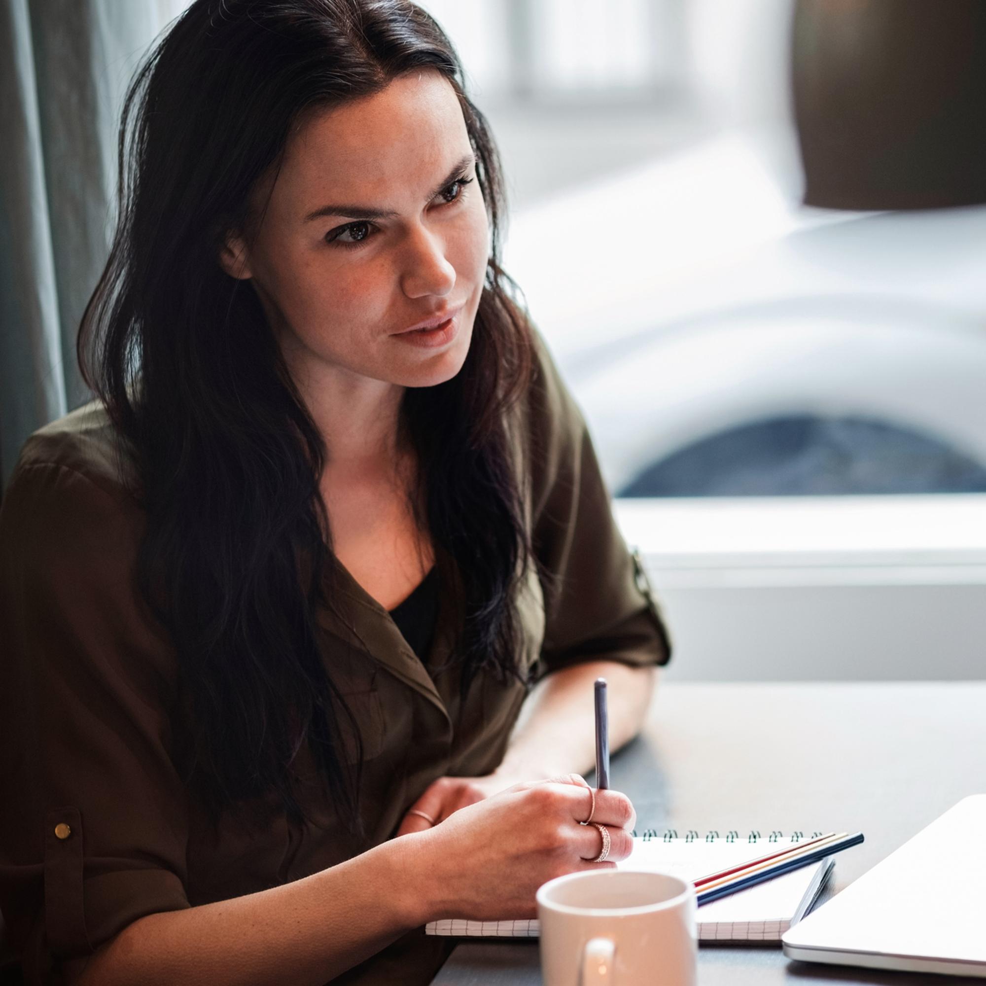 Kvinna med mörkt hår skriver i anteckningsblock vid ett bord, bredvid en kaffekopp och en bärbar dator