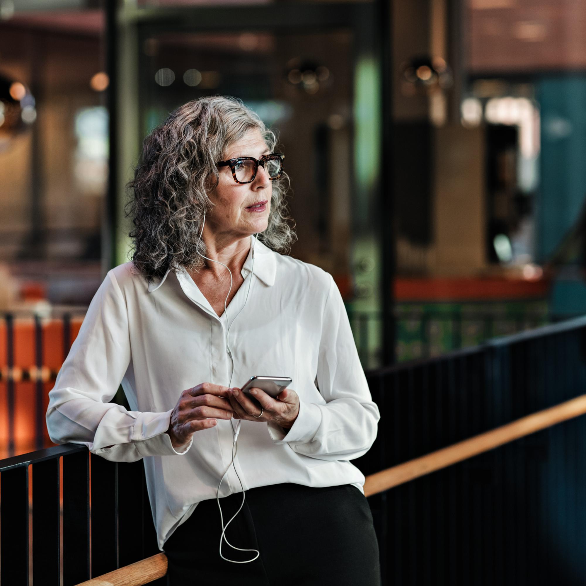 Äldre kvinna med grått hår och glasögon står inomhus, klädd i vit skjorta, och håller en smartphone med hörlurar i handen. Hon verkar fundersam och avkopplad