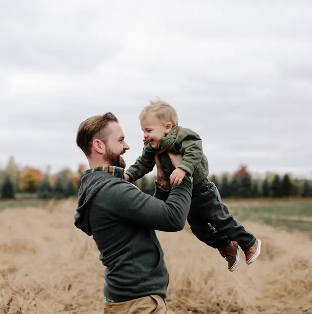 Man lyfter upp ett leende barn i en höstlig utomhusmiljö med träd i bakgrunden