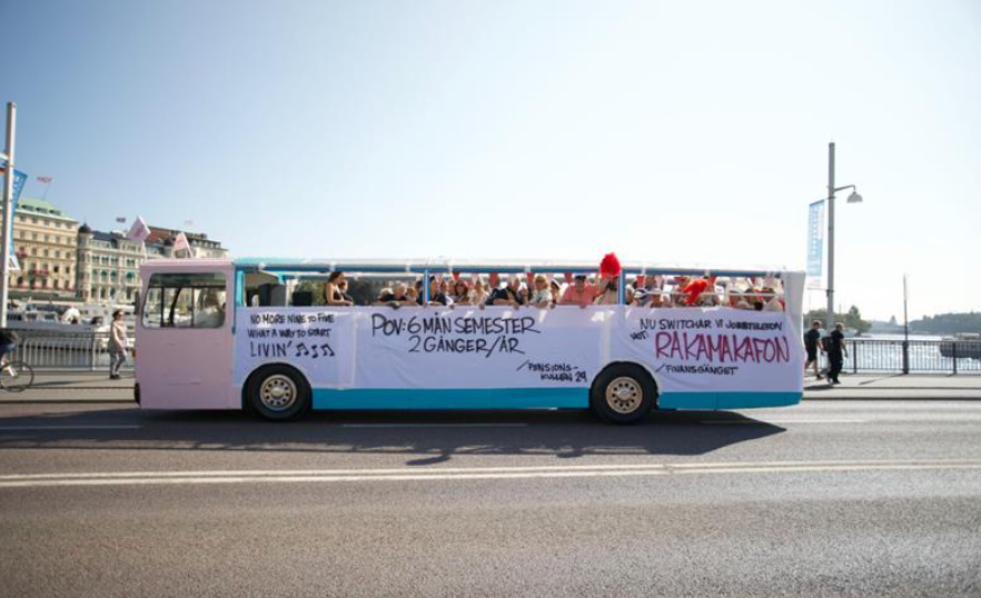 En öppen buss kör genom en stad med en stor banderoll som marknadsför sex månaders semester två gånger om året. Bakgrunden visar byggnader och en vattenutsikt