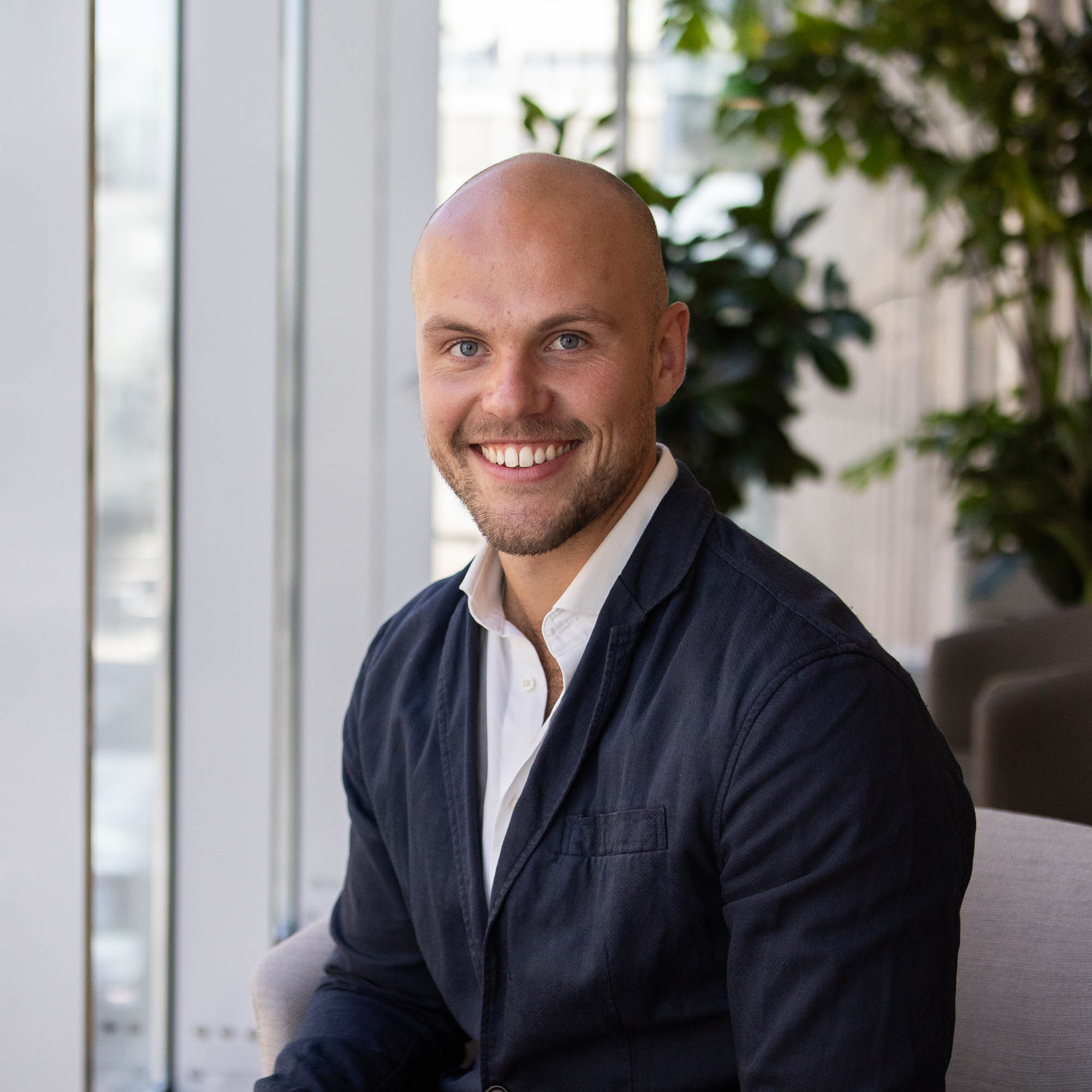 Leende man i mörk kavaj sitter inomhus med gröna växter i bakgrunden