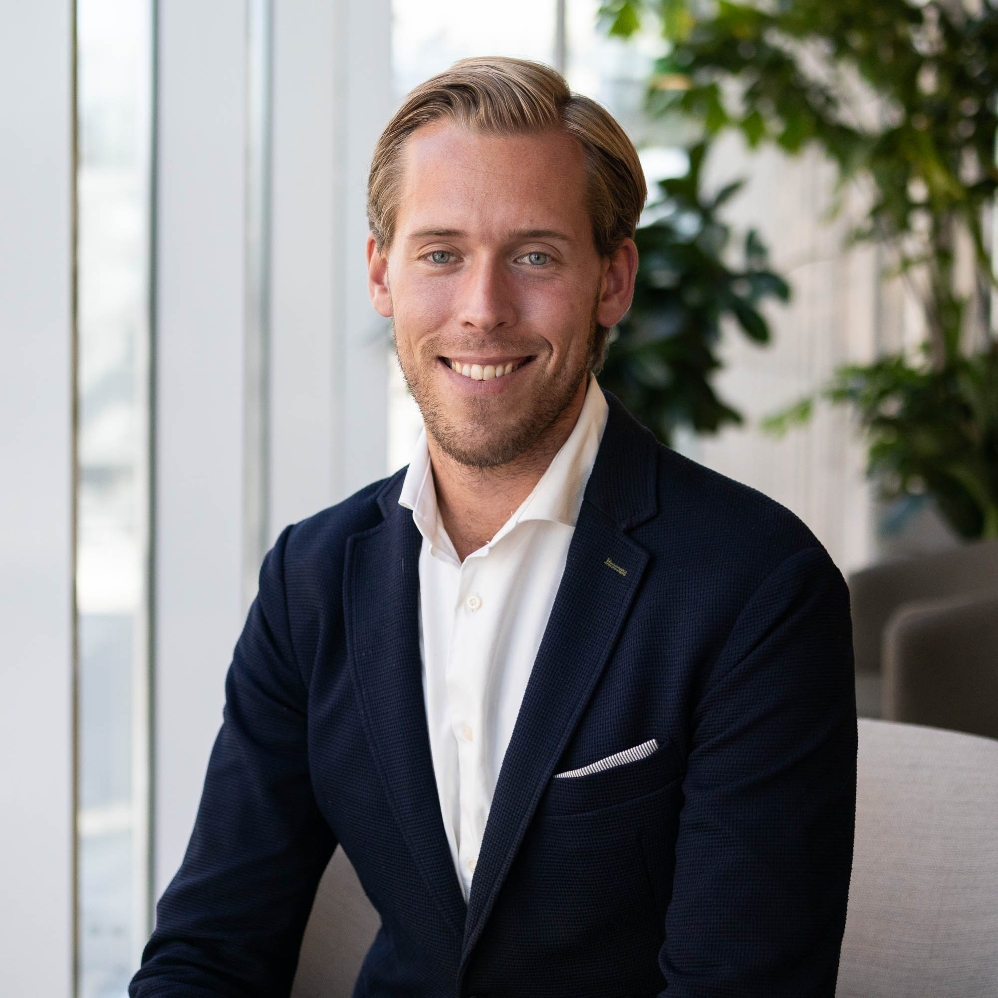 Man i kostym och vit skjorta som ler, sitter i en modern kontorsmiljö med gröna växter i bakgrunden