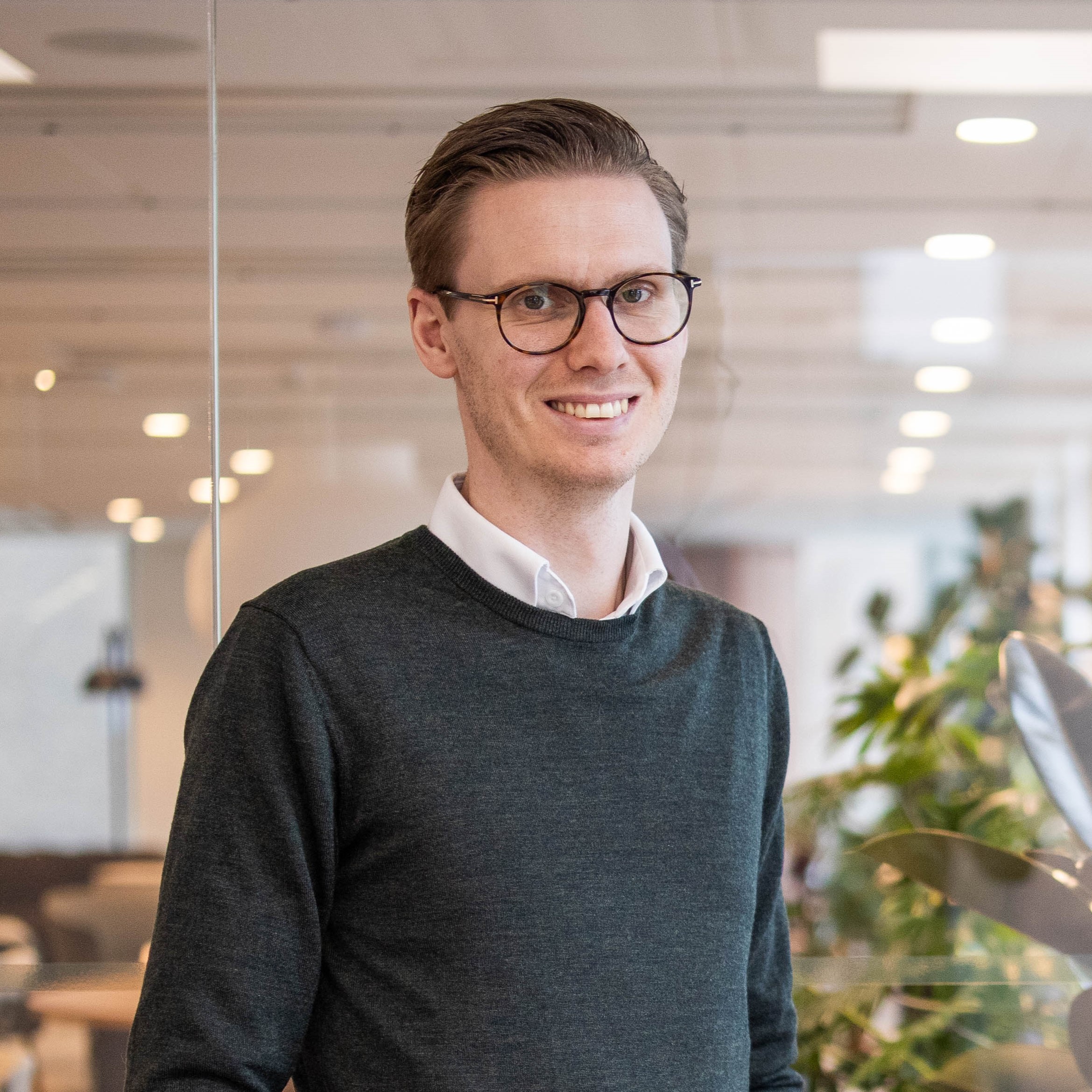 Man i glasögon och grön tröja i modernt kontor med växtlighet i bakgrunden