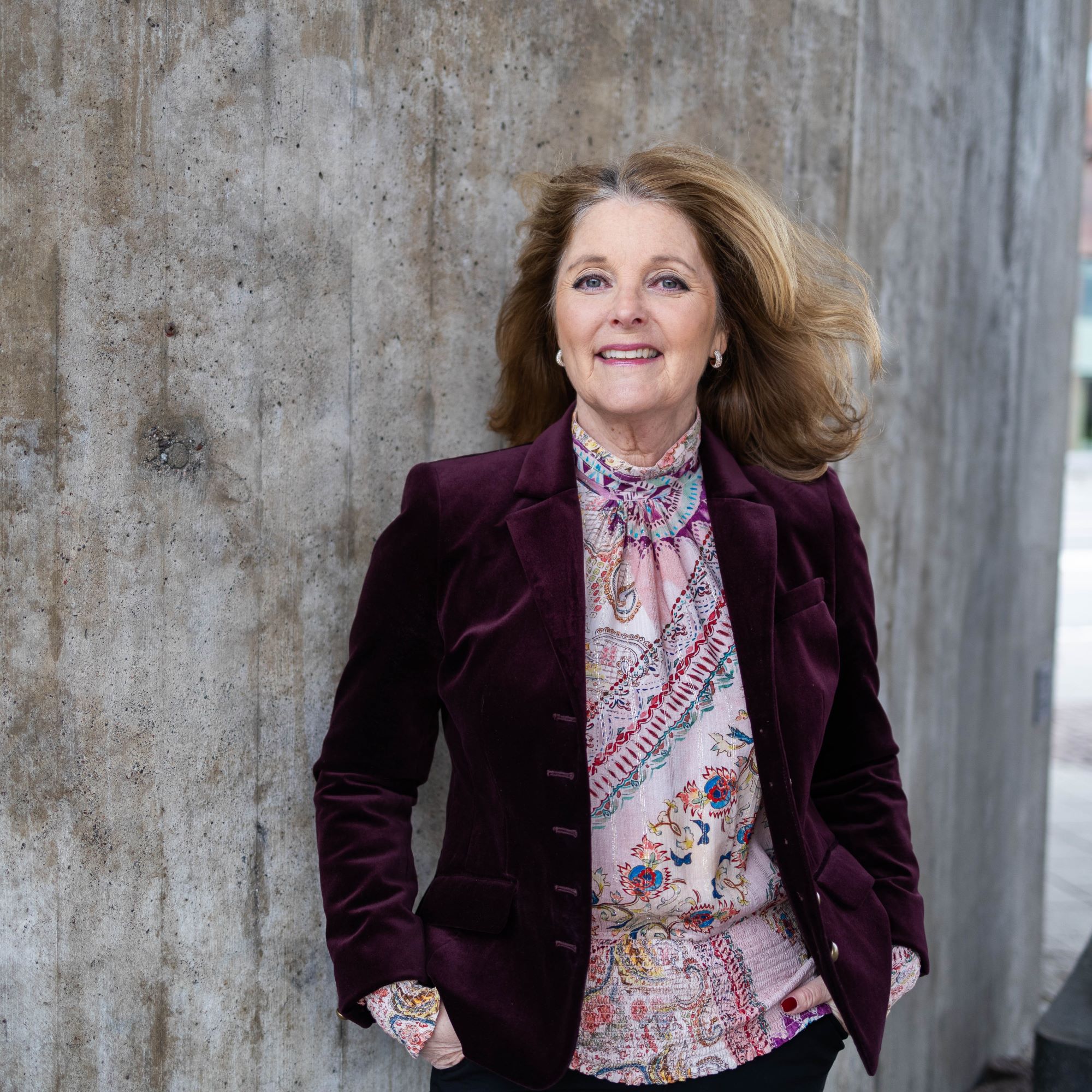 Kvinna i vinröd kavaj och mönstrad blus står vid betongvägg utomhus, ler mot kameran