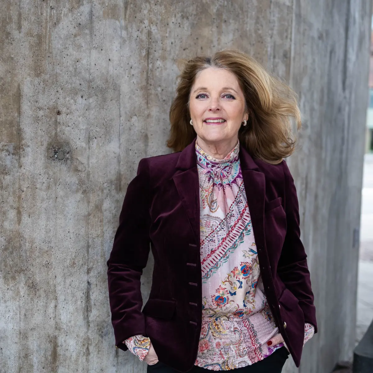 Kvinna i vinröd kavaj och mönstrad blus står vid betongvägg utomhus, ler mot kameran