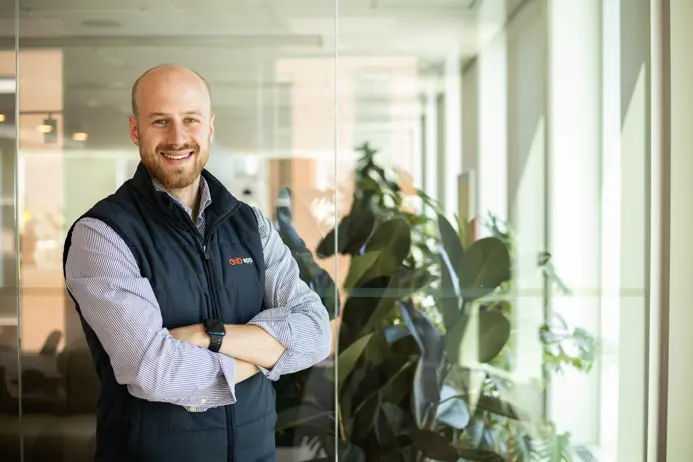 Man i kontorsmiljö med glasväggar, bär svart väst och skjorta, leende med armarna i kors, gröna växter i bakgrunden