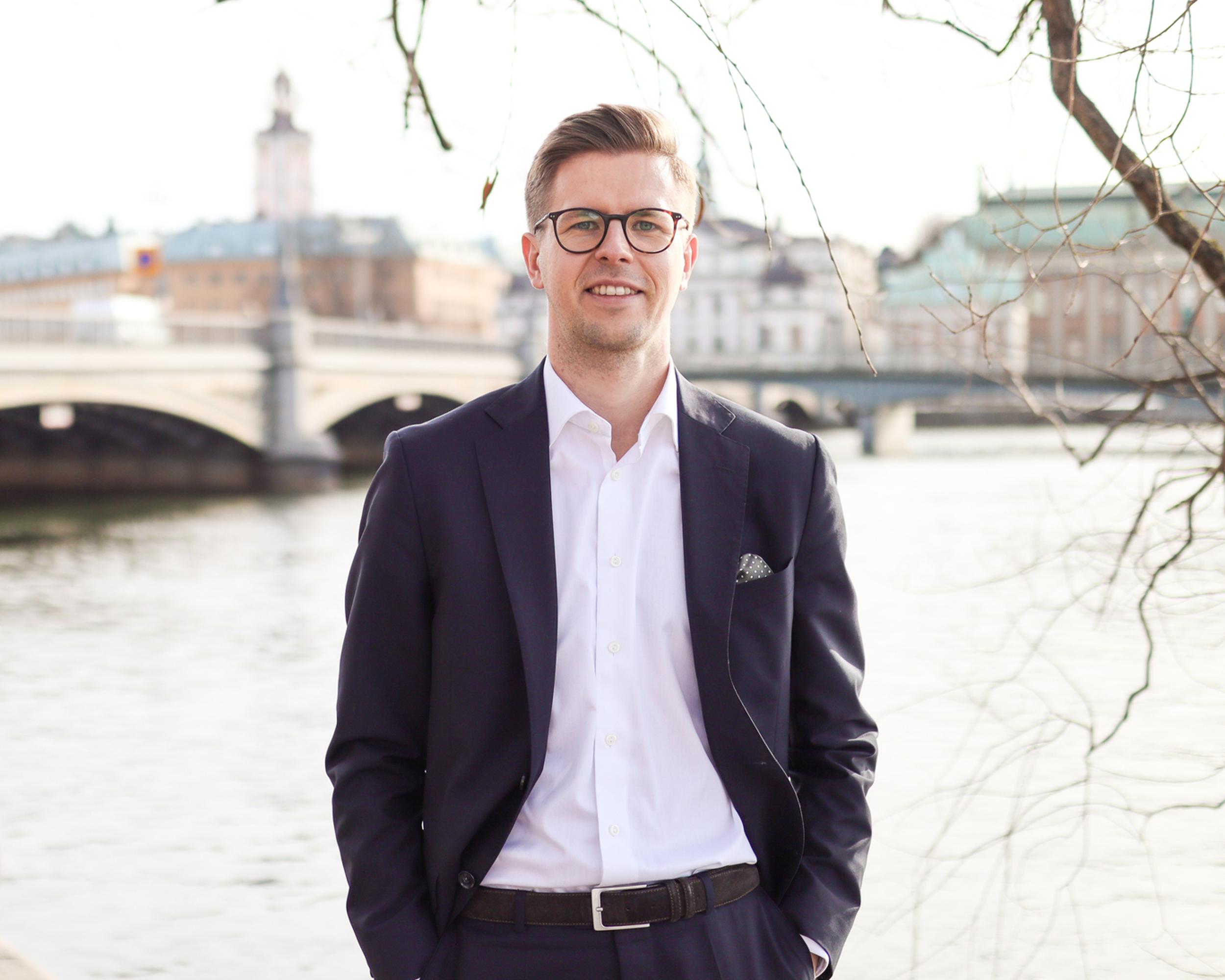 Man i kostym står framför vattnet med en bro och stadsbyggnader i bakgrunden, en solig dag i Stockholm