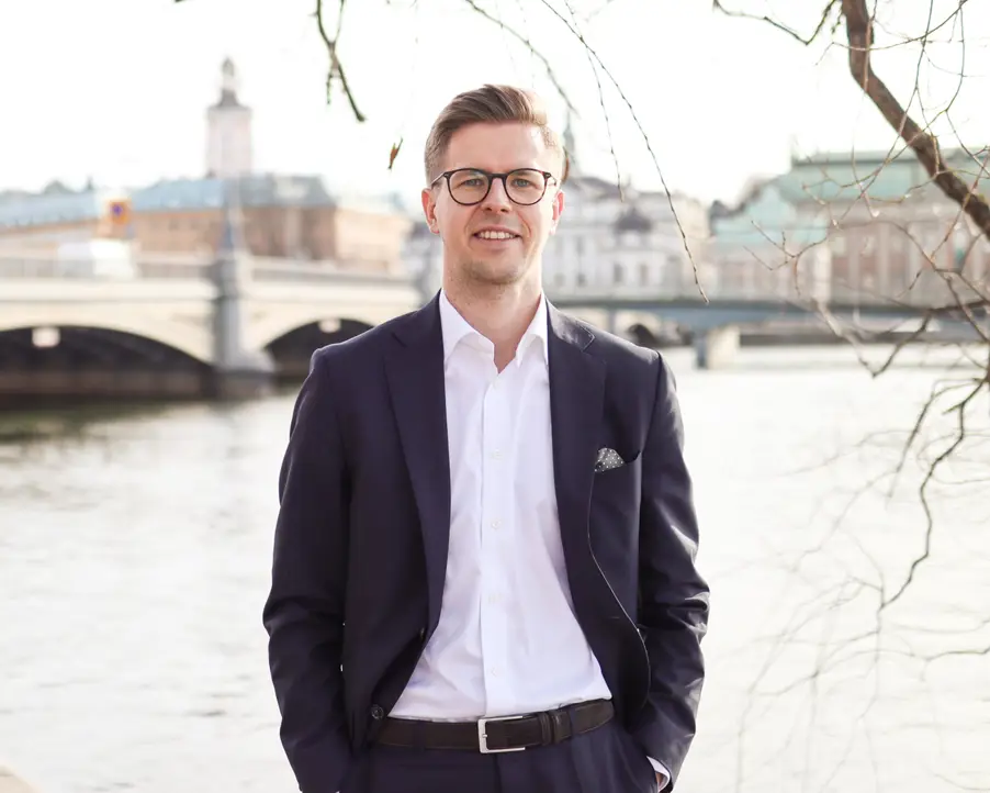 Man i kostym står framför vattnet med en bro och stadsbyggnader i bakgrunden, en solig dag i Stockholm
