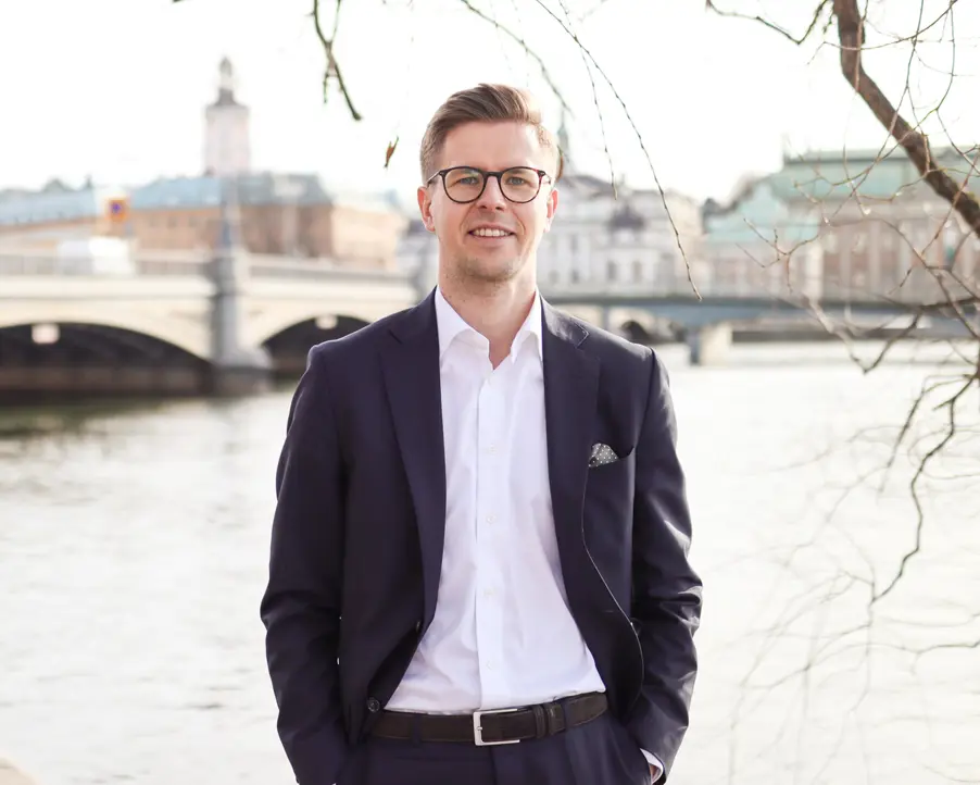 Man i kostym står framför vattnet med en bro och stadsbyggnader i bakgrunden, en solig dag i Stockholm