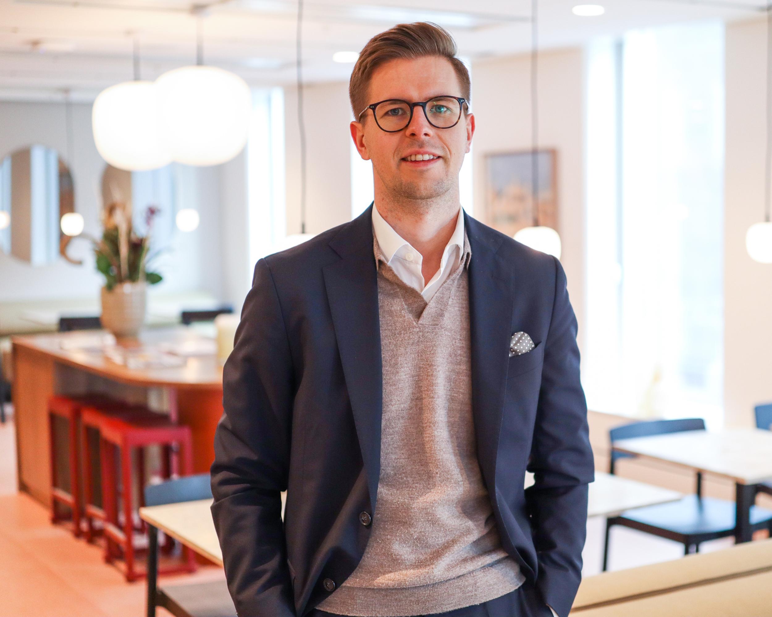 Man i kostym och glasögon i modernt kontorsmiljö med bord och stolar i bakgrunden