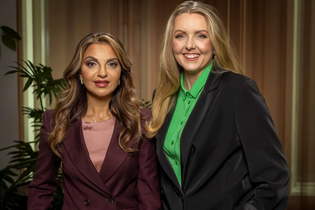 Två kvinnor i affärskläder står bredvid varandra i en inomhusmiljö med växter i bakgrunden