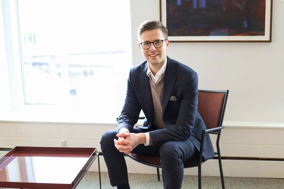 Man i kostym som sitter på en stol i ett modernt kontor med ett leende, stora fönster i bakgrunden