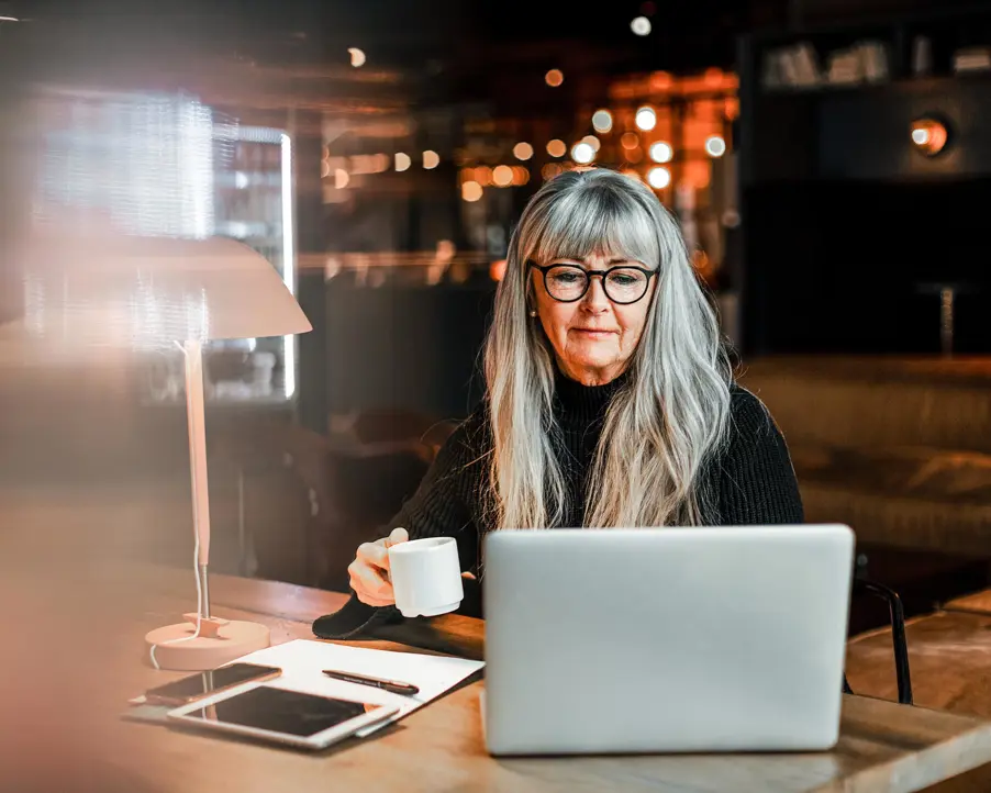 Äldre kvinna med långt grått hår och glasögon dricker kaffe medan hon arbetar på en laptop i ett mysigt café