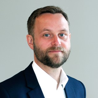 Man i kostym med mörkt hår och skägg står mot en grå bakgrund