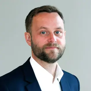 Man i kostym med mörkt hår och skägg står mot en grå bakgrund