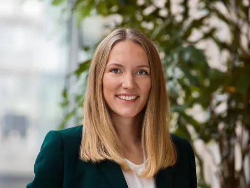 Person med långt blont hår och grön kavaj, ler framför en bakgrund av gröna växter och glasfönster