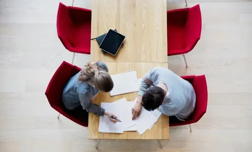 Två personer diskuterar över ett bord med dokument och surfplatta i en kontorsmiljö