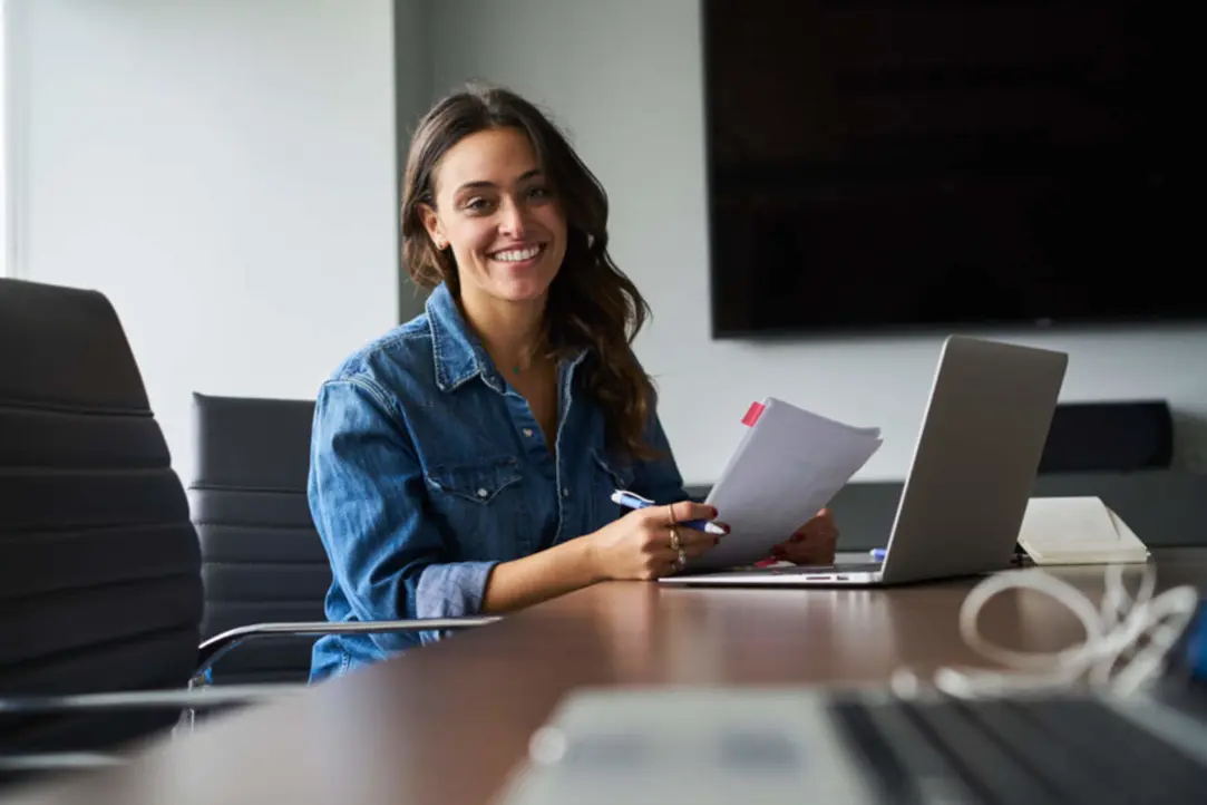 Kvinna i jeansskjorta arbetar vid en bärbar dator, håller dokument och ler in i kameran, i ett modernt kontorsmiljö