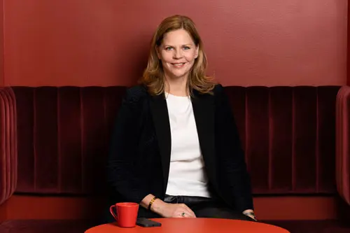 Kvinna i svart kavaj sitter på röd soffa med kaffekopp på bordet