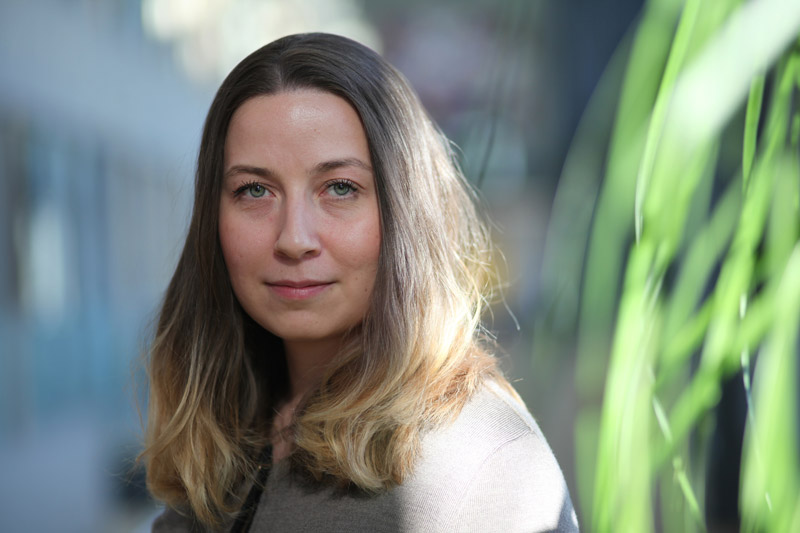 Kvinna med långt brunt hår står inomhus med grön växt i förgrunden