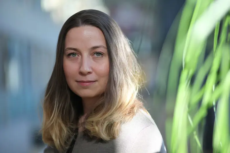 Kvinna med långt brunt hår står inomhus med grön växt i förgrunden