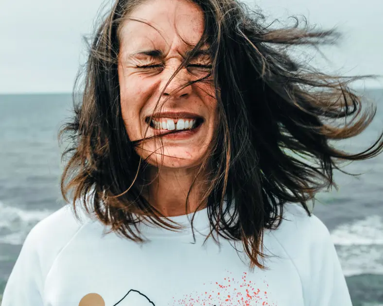 Kvinna med blåsigt hår vid havet, ler med stängda ögon i vinden