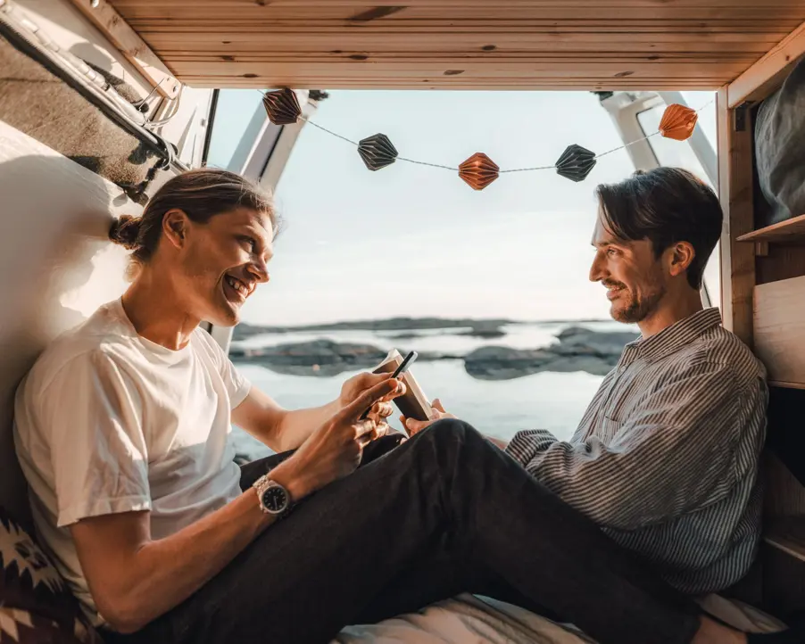 Två glada män sitter i en campervan vid havet under en sommarkväll, skrattandes och tittandes på en mobiltelefon