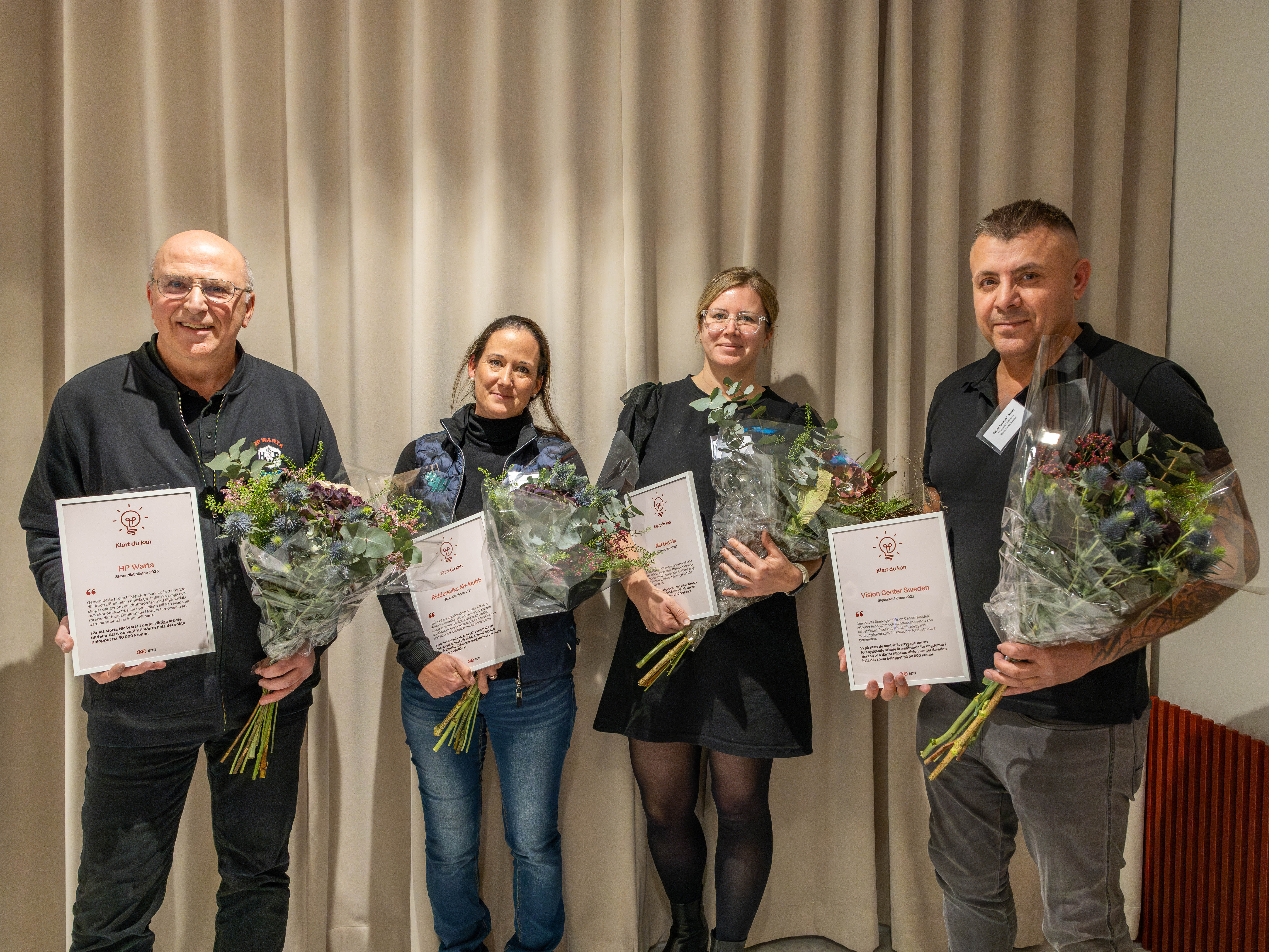 Fyra personer håller blombuketter och certifikat framför ett draperi vid en utmärkelseceremoni