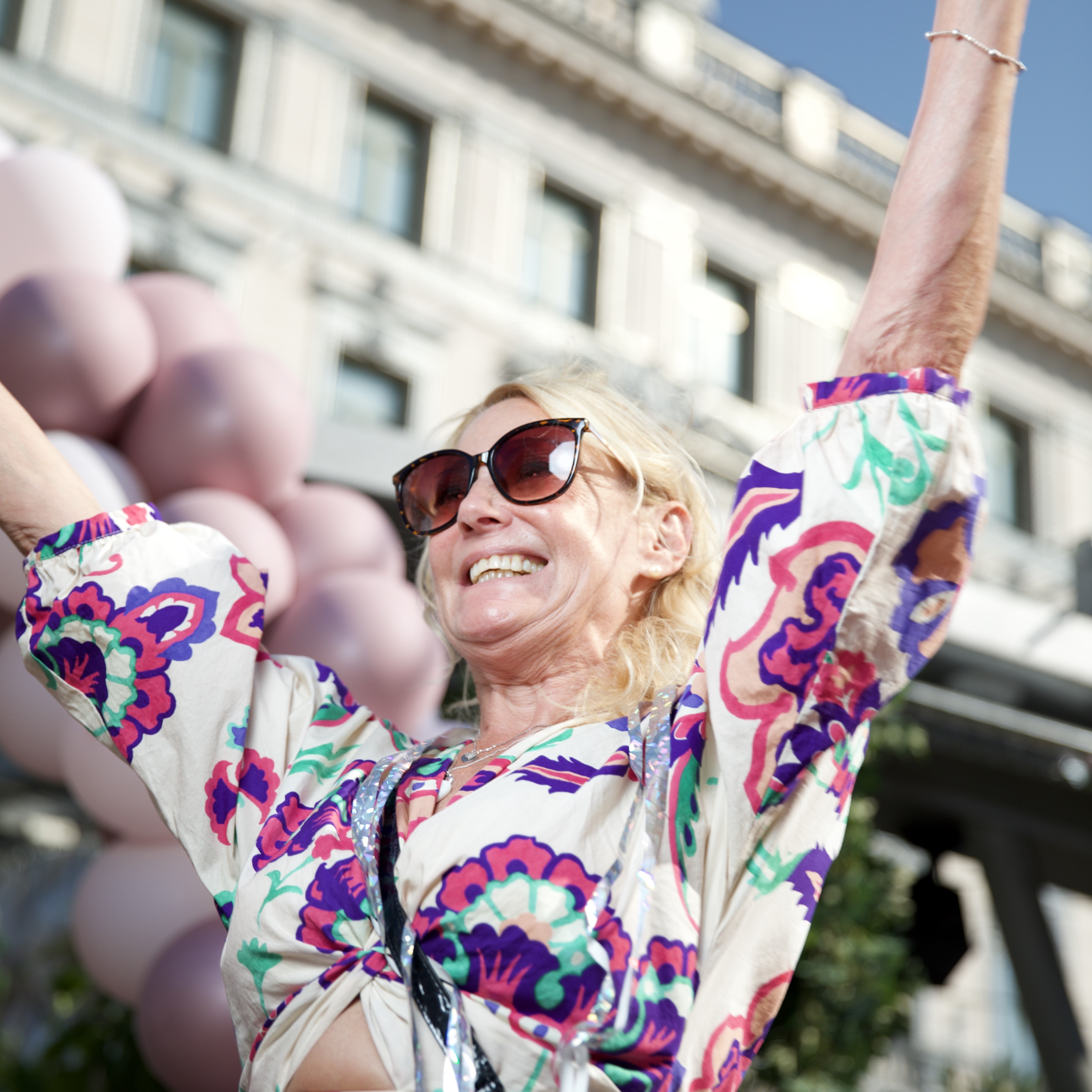 Kvinna i färgglad klänning och solglasögon jublar utomhus framför ett hus med ballonger i bakgrunden