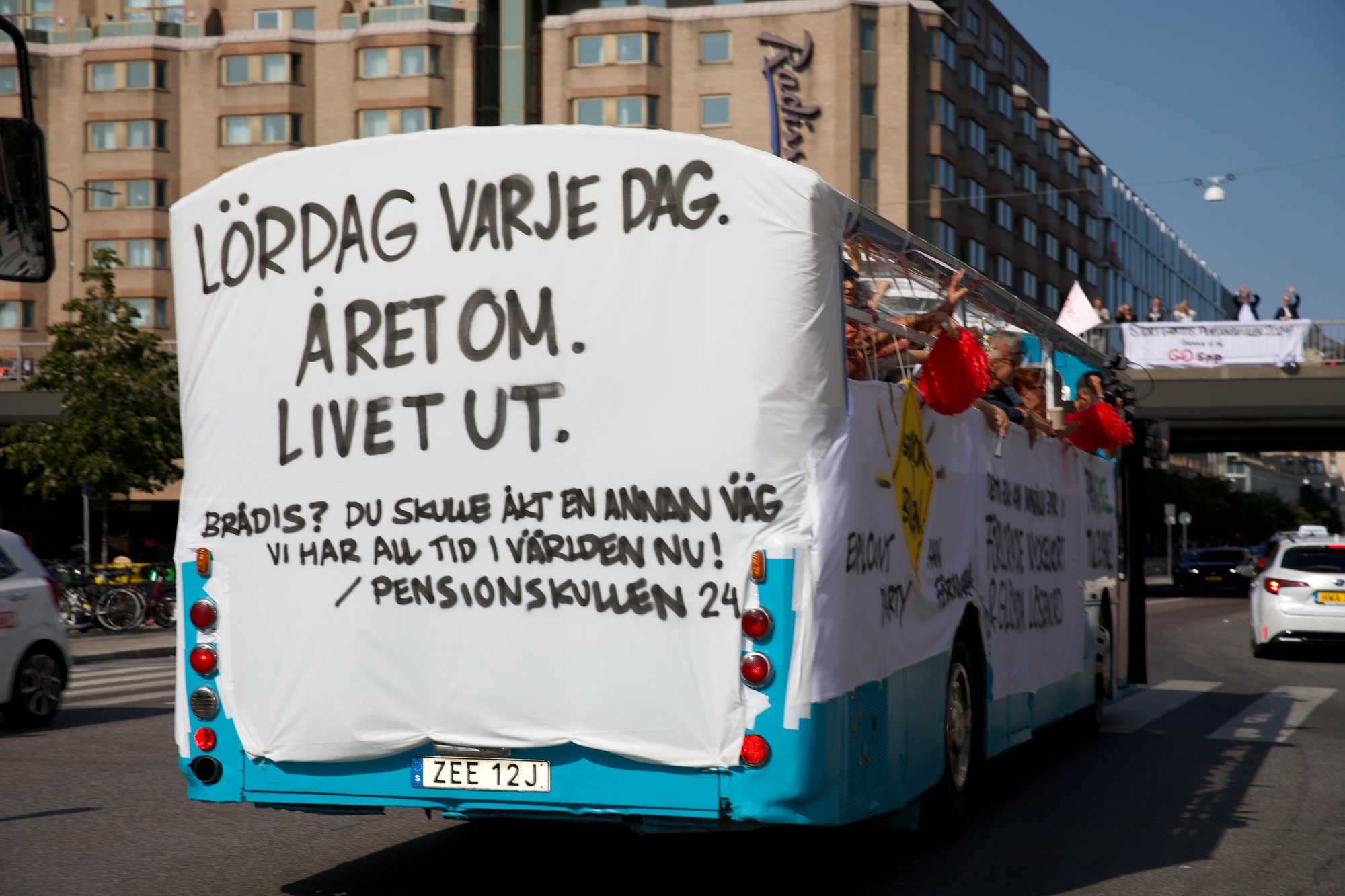 Karnevalsbuss dekorerad med texten "Lördag varje dag. Året om. Livet ut." som kör genom en stadsgata i Sverige