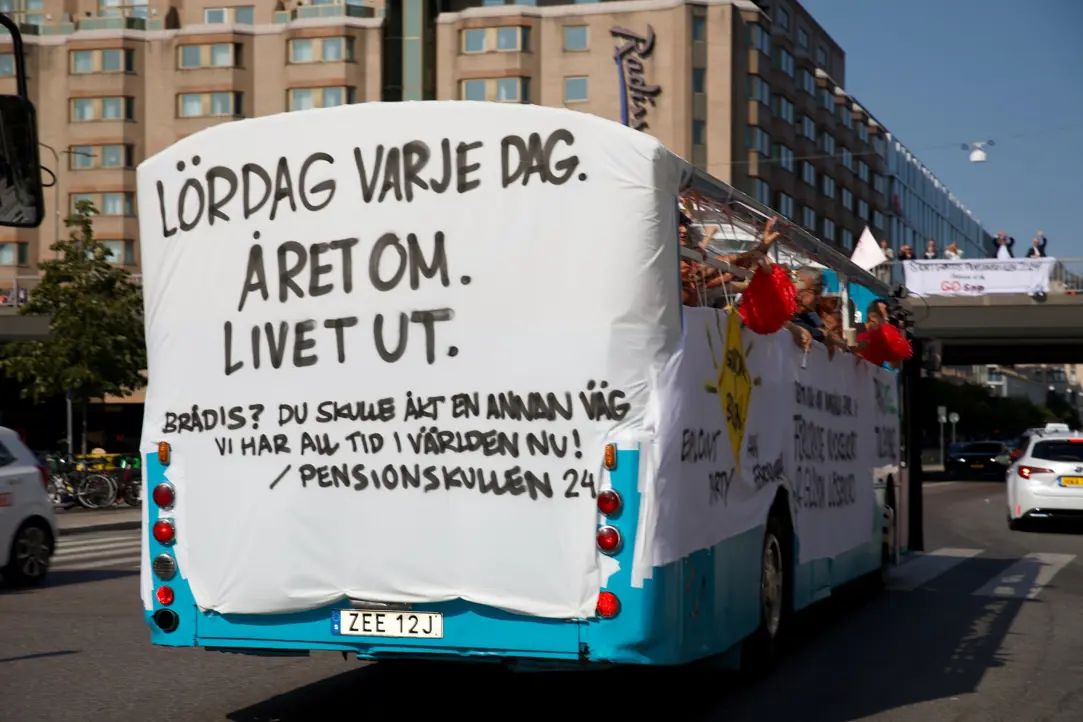 Karnevalsbuss dekorerad med texten "Lördag varje dag. Året om. Livet ut." som kör genom en stadsgata i Sverige