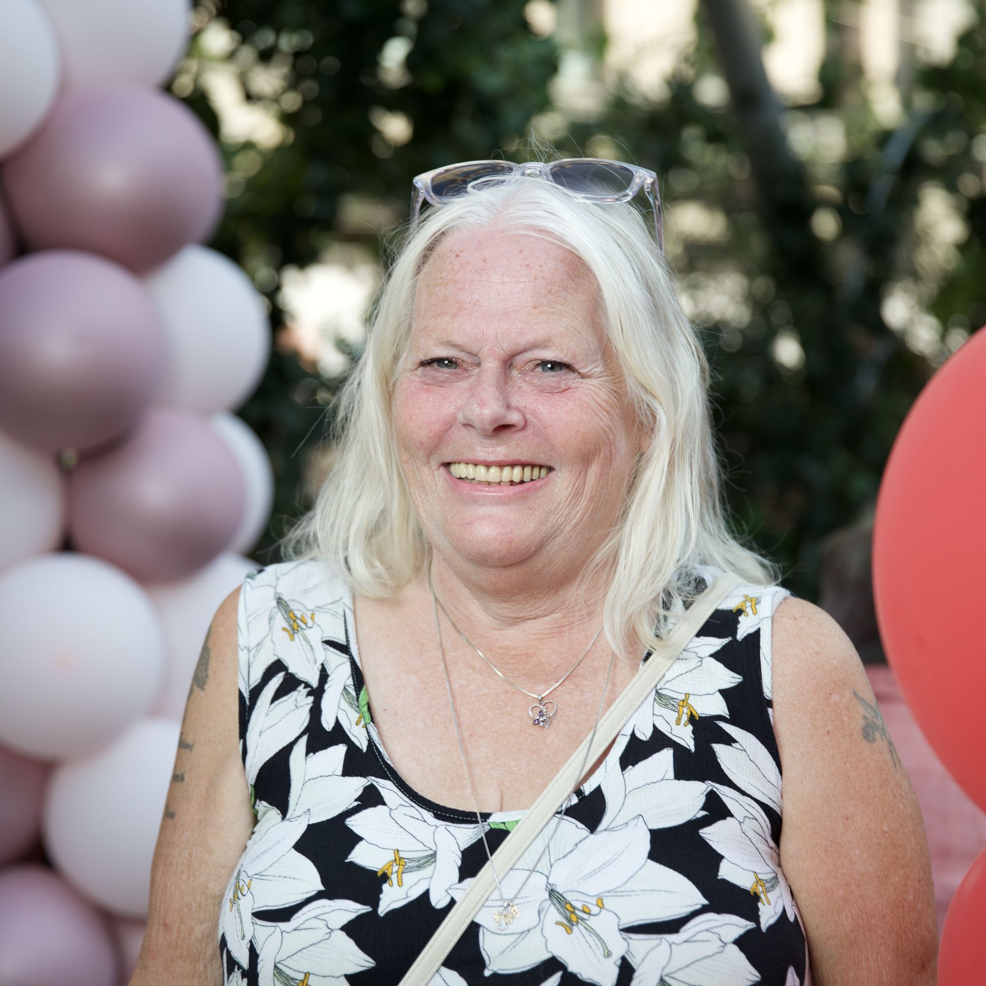 Kvinna med långt vitt hår och blommig klänning står leende framför en bakgrund av pastellfärgade ballonger utomhus